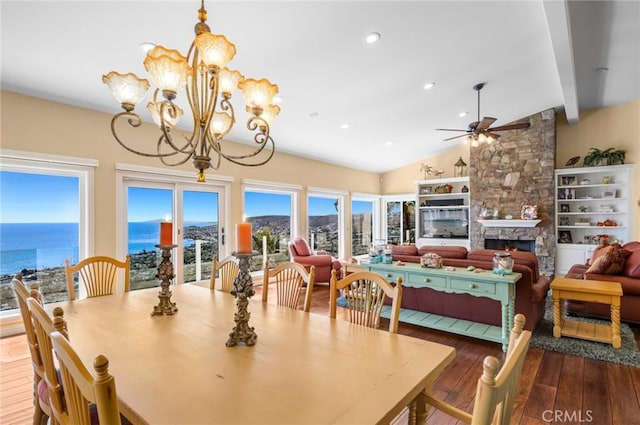 dining area featuring ceiling fan with notable chandelier, dark hardwood / wood-style flooring, a stone fireplace, lofted ceiling with beams, and a water view