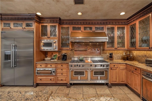 kitchen with light tile patterned flooring, built in appliances, light stone countertops, and decorative backsplash