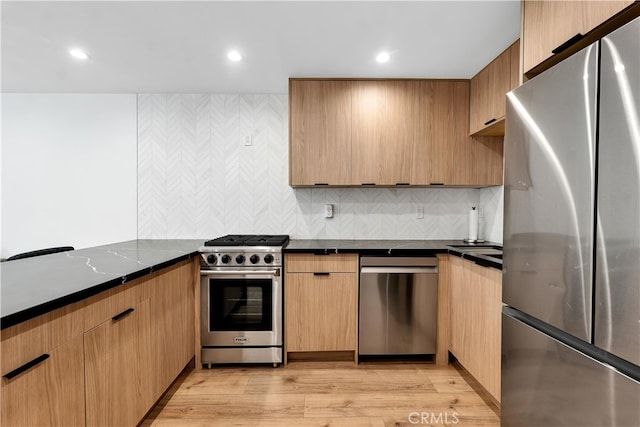 kitchen with light brown cabinetry, light hardwood / wood-style flooring, dark stone countertops, appliances with stainless steel finishes, and decorative backsplash