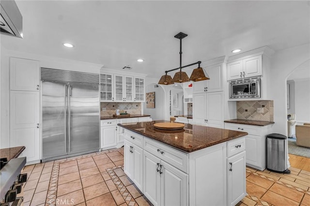 kitchen featuring white cabinetry, hanging light fixtures, a kitchen island, built in appliances, and dark stone counters