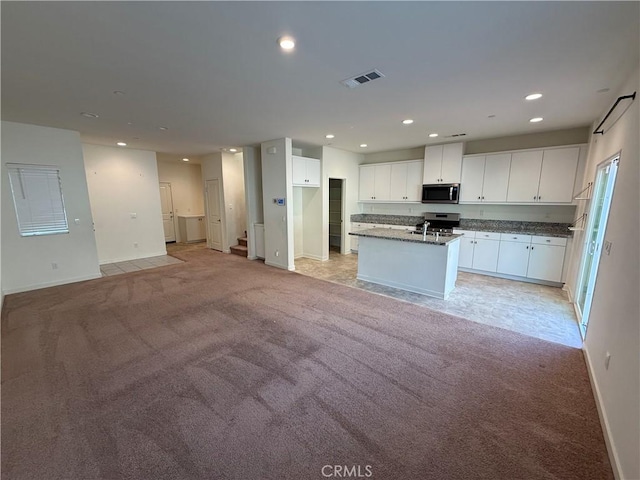 kitchen with appliances with stainless steel finishes, white cabinetry, dark stone countertops, a center island with sink, and light colored carpet