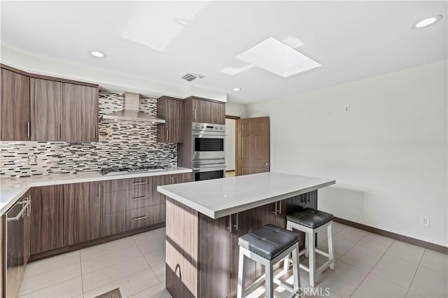 kitchen with appliances with stainless steel finishes, wall chimney exhaust hood, a kitchen island, tasteful backsplash, and a breakfast bar area