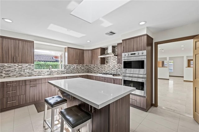 kitchen featuring wall chimney exhaust hood, a kitchen island, a kitchen bar, double oven, and white gas cooktop