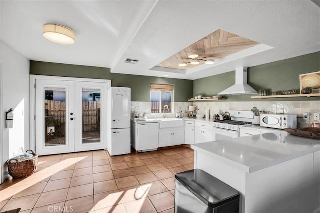 kitchen with white appliances, wall chimney exhaust hood, white cabinetry, sink, and a raised ceiling