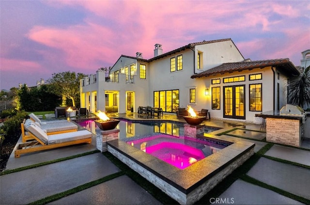 back house at dusk with an in ground hot tub, a balcony, a patio, and a fire pit