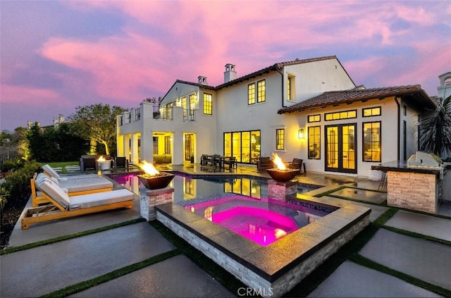 back of house at dusk featuring stucco siding, an outdoor kitchen, an outdoor fire pit, a patio area, and an in ground hot tub