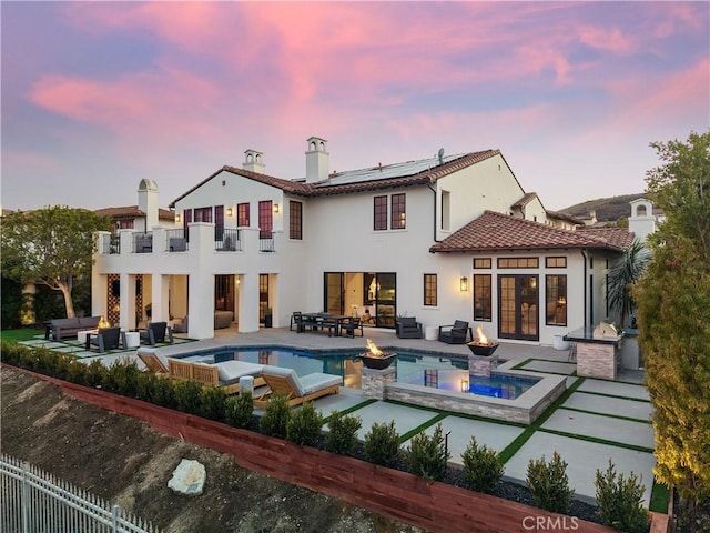 back house at dusk with solar panels, a balcony, a swimming pool with hot tub, a patio, and an outdoor living space with a fire pit