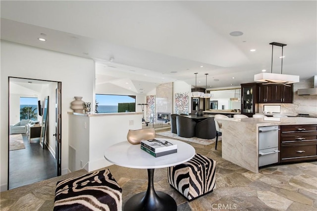 interior space with hanging light fixtures, wall chimney exhaust hood, light stone counters, decorative backsplash, and dark brown cabinetry