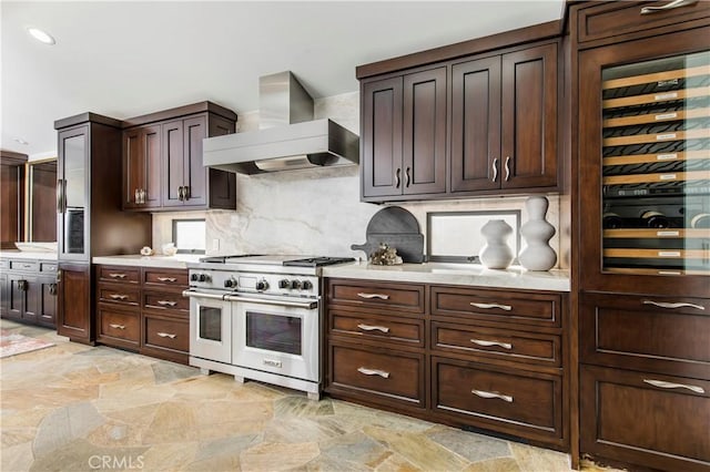 kitchen featuring wall chimney range hood, dark brown cabinetry, backsplash, beverage cooler, and range with two ovens