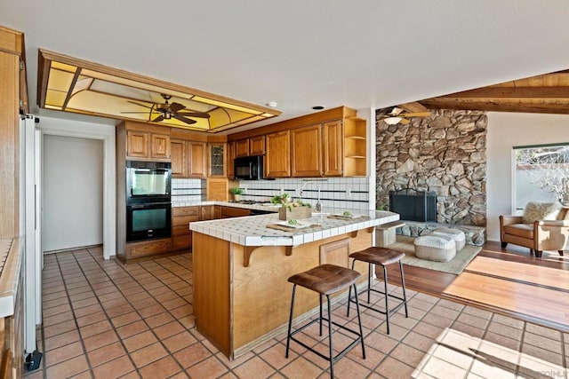 kitchen with a breakfast bar, ceiling fan, tile counters, black appliances, and kitchen peninsula