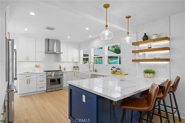 kitchen with appliances with stainless steel finishes, white cabinetry, wall chimney range hood, decorative light fixtures, and kitchen peninsula