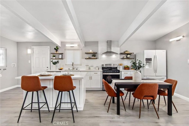 kitchen featuring white fridge with ice dispenser, wall chimney exhaust hood, a kitchen island, white cabinetry, and gas range