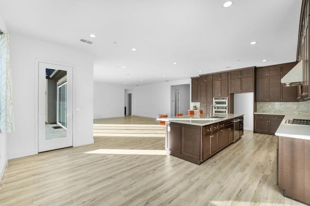 kitchen featuring tasteful backsplash, a kitchen island with sink, stainless steel gas cooktop, dark brown cabinetry, and light hardwood / wood-style flooring