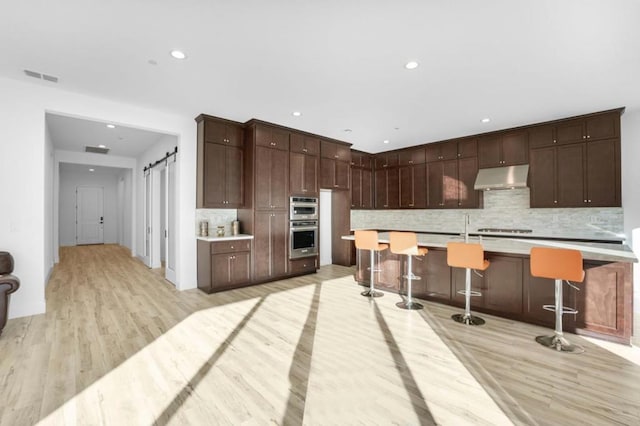 kitchen featuring a kitchen breakfast bar, dark brown cabinetry, a barn door, a center island with sink, and light wood-type flooring