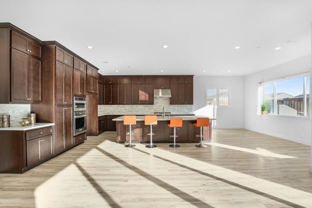 kitchen featuring a kitchen bar, decorative backsplash, a kitchen island with sink, dark brown cabinetry, and light wood-type flooring