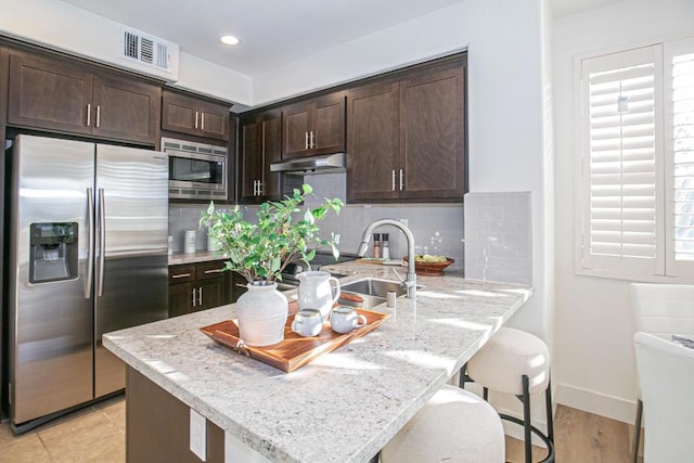 kitchen with dark brown cabinetry, appliances with stainless steel finishes, a kitchen bar, and light stone countertops