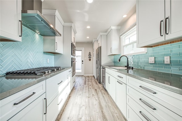 kitchen featuring white cabinets, wall chimney range hood, stainless steel appliances, sink, and light hardwood / wood-style flooring