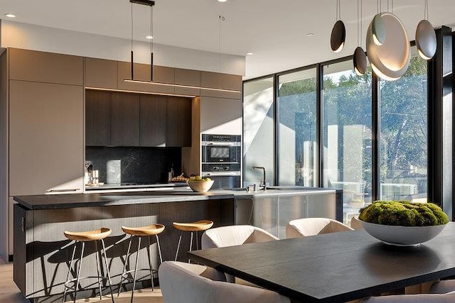 kitchen featuring double oven, hanging light fixtures, a kitchen bar, a wall of windows, and decorative backsplash