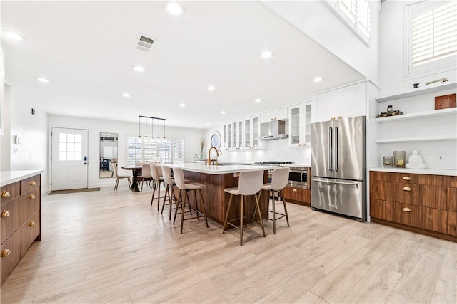 kitchen featuring white cabinets, stainless steel appliances, an island with sink, ventilation hood, and a breakfast bar area