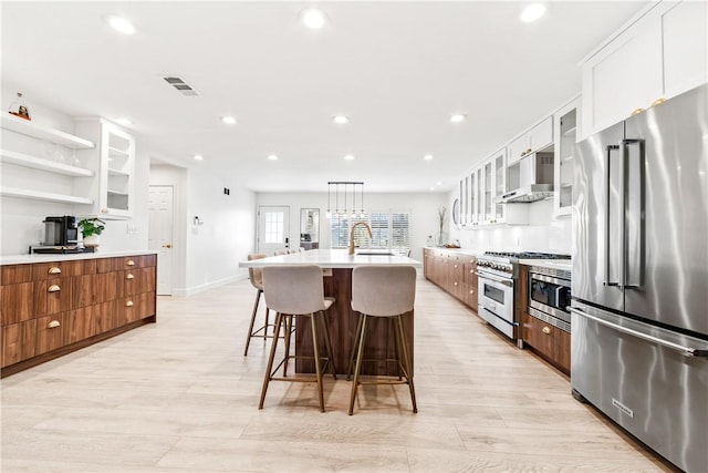 kitchen featuring premium appliances, a kitchen island with sink, white cabinets, pendant lighting, and wall chimney exhaust hood