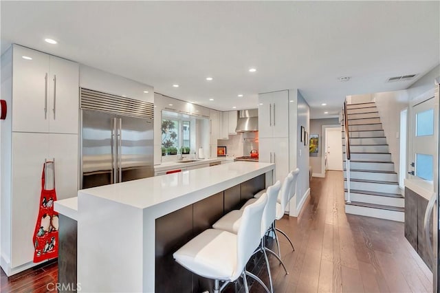 kitchen featuring white cabinets, built in refrigerator, wall chimney range hood, a kitchen island, and a breakfast bar area