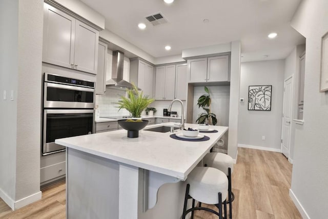 kitchen with gray cabinets, double oven, sink, a kitchen island with sink, and wall chimney range hood