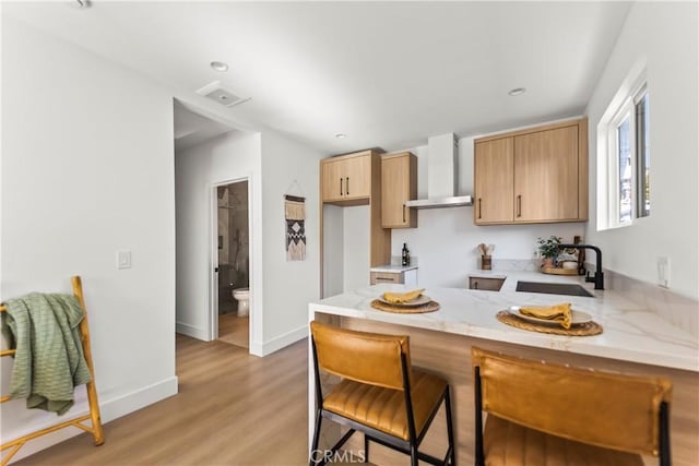 kitchen featuring wall chimney range hood, sink, light brown cabinetry, kitchen peninsula, and light wood-type flooring