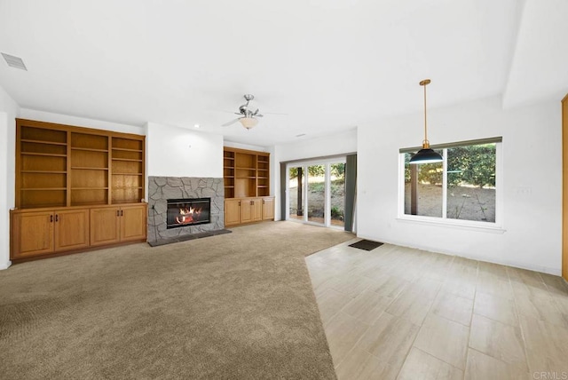 unfurnished living room featuring built in features, ceiling fan, a stone fireplace, and light colored carpet