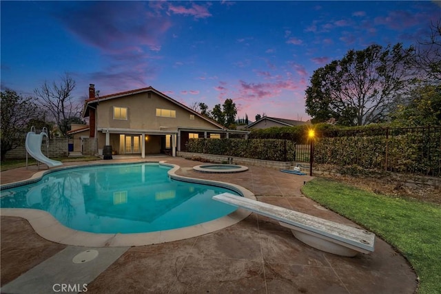 pool at dusk featuring an in ground hot tub, a diving board, a water slide, and a patio