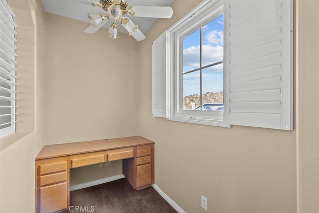 unfurnished office featuring ceiling fan, built in desk, dark carpet, and a wealth of natural light