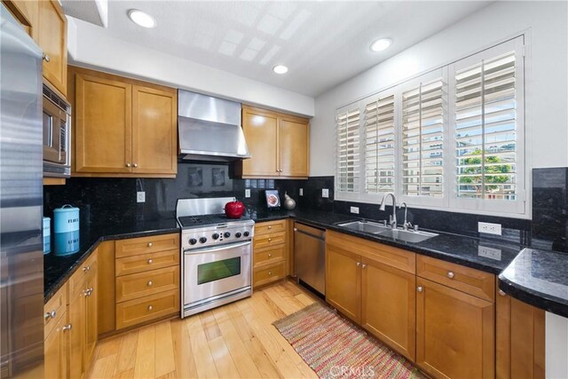 kitchen featuring wall chimney exhaust hood, sink, dark stone countertops, light hardwood / wood-style floors, and stainless steel appliances