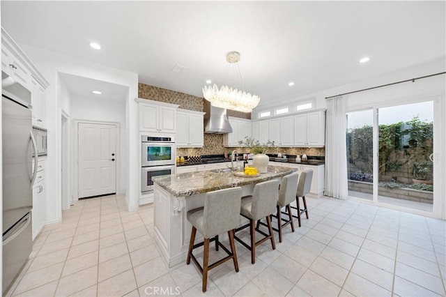 kitchen with a kitchen island with sink, stainless steel appliances, white cabinets, wall chimney range hood, and light stone countertops