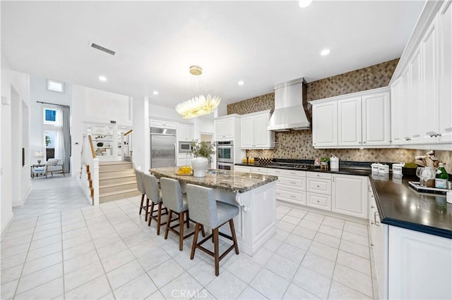 kitchen with visible vents, white cabinets, a kitchen island, built in appliances, and wall chimney range hood