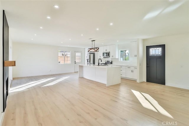 kitchen with white cabinets, decorative light fixtures, stainless steel appliances, an island with sink, and light hardwood / wood-style floors