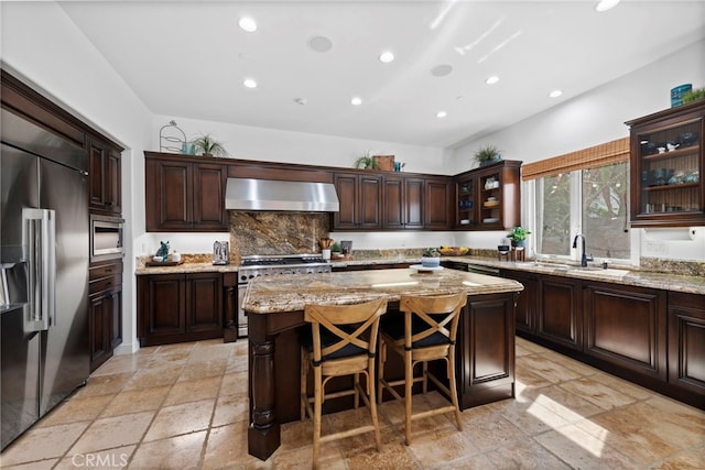 kitchen featuring a kitchen island, wall chimney range hood, built in appliances, sink, and light stone counters