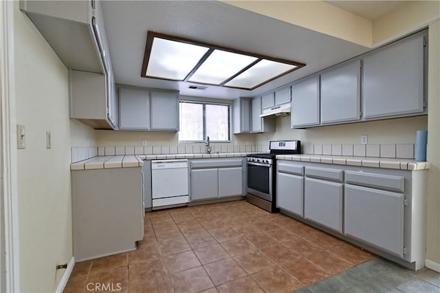 kitchen with gray cabinets, tile countertops, white dishwasher, light tile patterned floors, and stainless steel range with gas stovetop