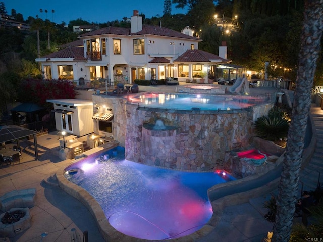 pool at night featuring a fire pit, exterior kitchen, a patio, and a water slide