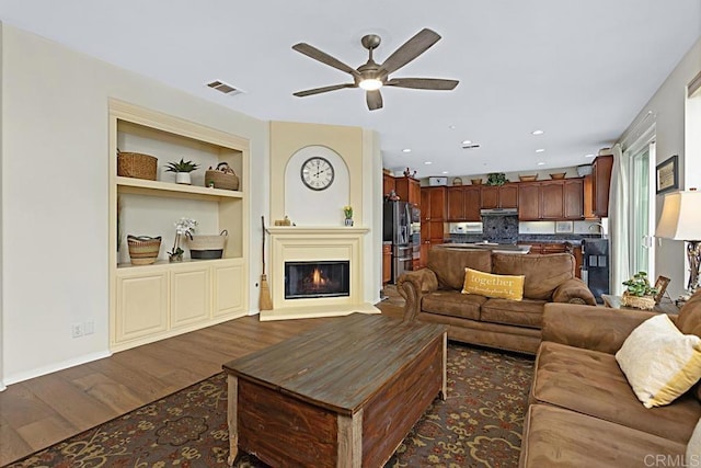 living room with built in shelves, ceiling fan, and dark wood-type flooring