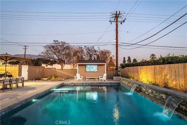 pool at dusk with pool water feature, an outbuilding, and a patio area