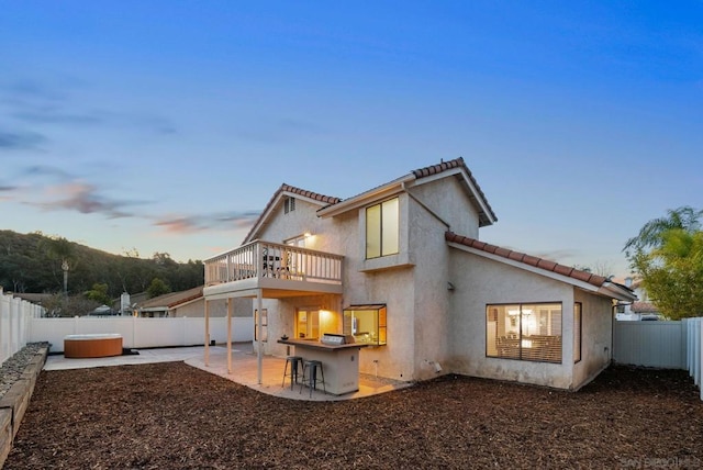 back house at dusk with a balcony, a patio area, exterior bar, and a hot tub