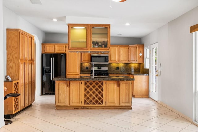 kitchen featuring decorative backsplash, dark stone countertops, an island with sink, stainless steel appliances, and light tile patterned flooring