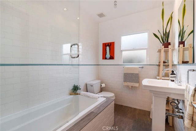 bathroom with wood-type flooring, tiled bath, tile walls, and toilet