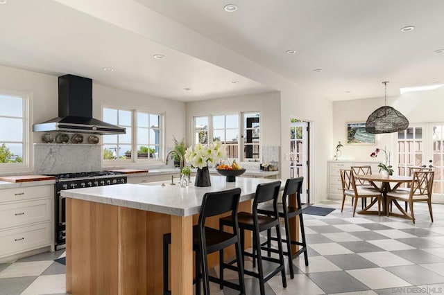 kitchen featuring wall chimney exhaust hood, decorative light fixtures, white cabinetry, a kitchen island with sink, and gas range