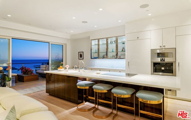 kitchen with light wood-type flooring, a water view, black electric stovetop, white cabinets, and a kitchen bar