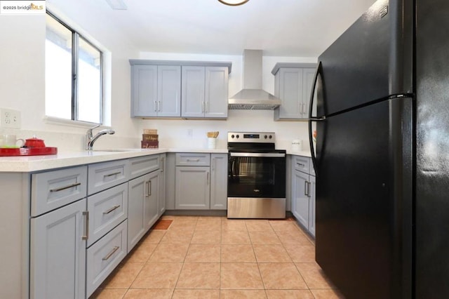 kitchen featuring black refrigerator, stainless steel electric range oven, light tile patterned floors, gray cabinetry, and wall chimney range hood