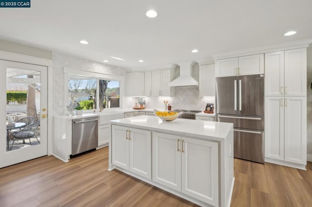 kitchen with appliances with stainless steel finishes, sink, white cabinets, wall chimney range hood, and light hardwood / wood-style flooring