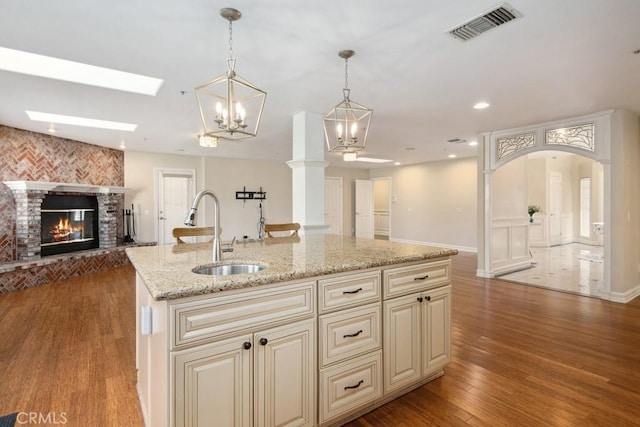 kitchen with an island with sink, sink, hanging light fixtures, light stone countertops, and cream cabinetry