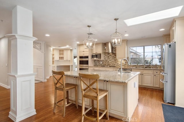 kitchen with appliances with stainless steel finishes, pendant lighting, a kitchen island with sink, light stone countertops, and light wood-type flooring