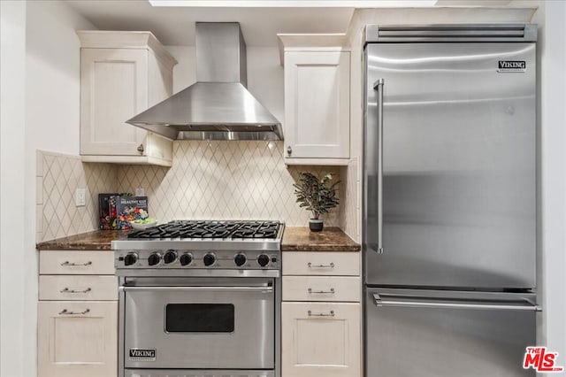 kitchen featuring tasteful backsplash, white cabinets, dark stone counters, high end appliances, and wall chimney exhaust hood