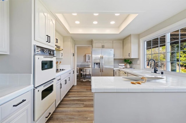 kitchen featuring stainless steel refrigerator with ice dispenser, black electric cooktop, white double oven, a raised ceiling, and kitchen peninsula